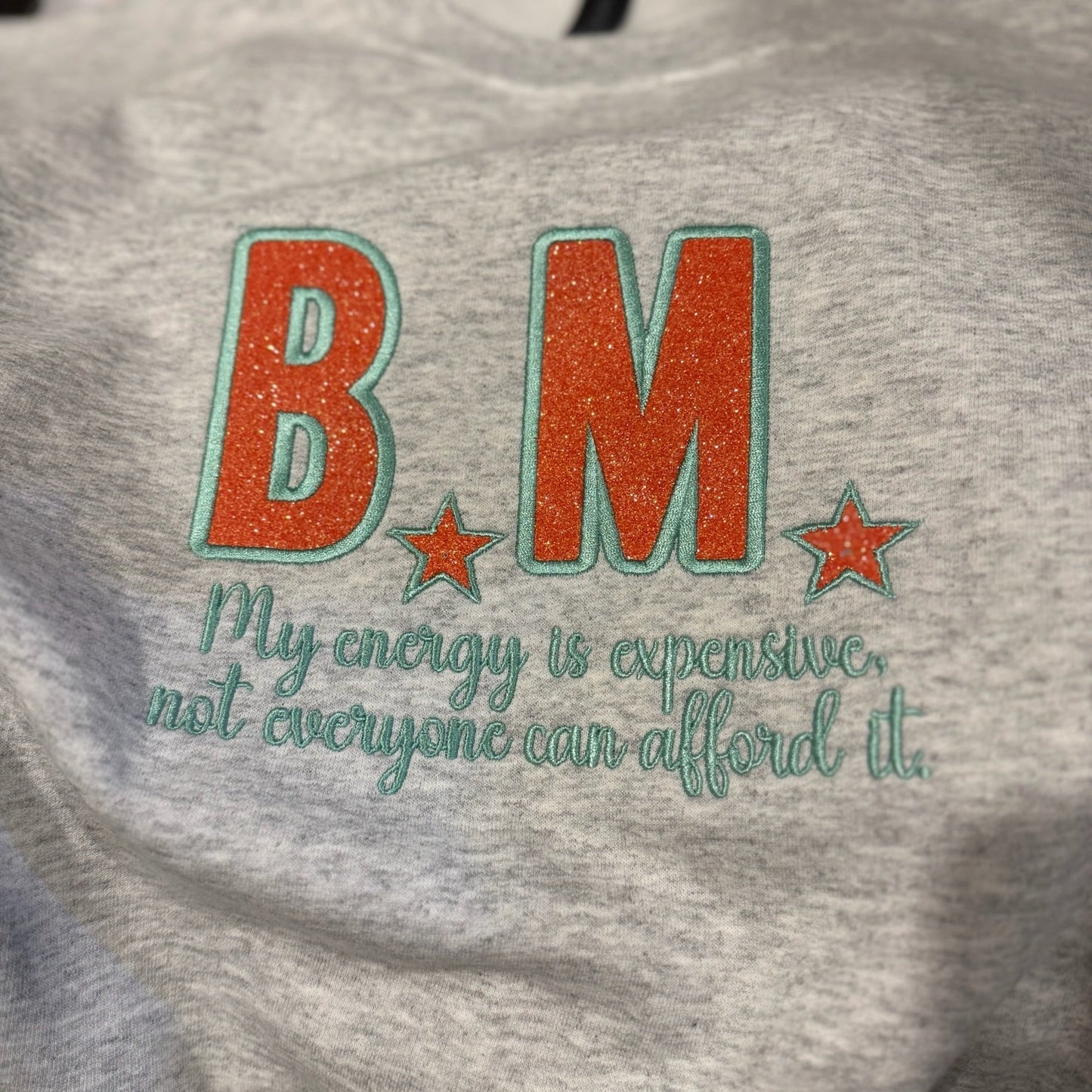Gray sweatshirt with orange applique initials and green text on a blurred background